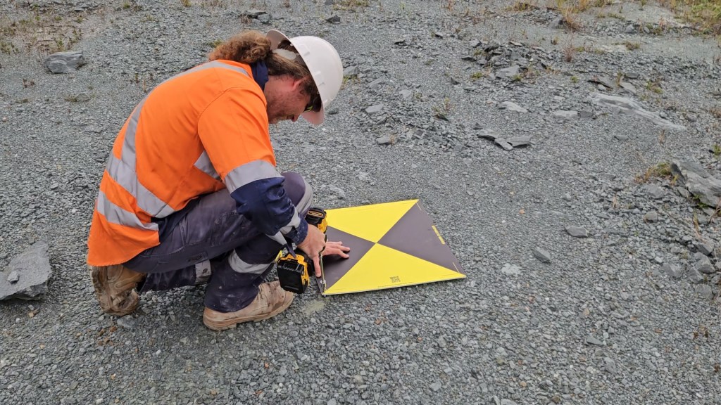 A worker in an orange safety vest and hard hat is kneeling on rocky ground, placing a yellow and black ground control point (GCP) marker with a triangular design.