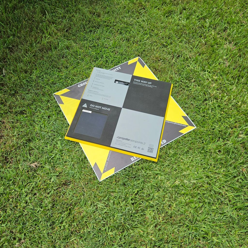 Aerial survey equipment, specifically a Propeller Aeropoint pad, positioned on grass, featuring visibility markings and instructional text.
