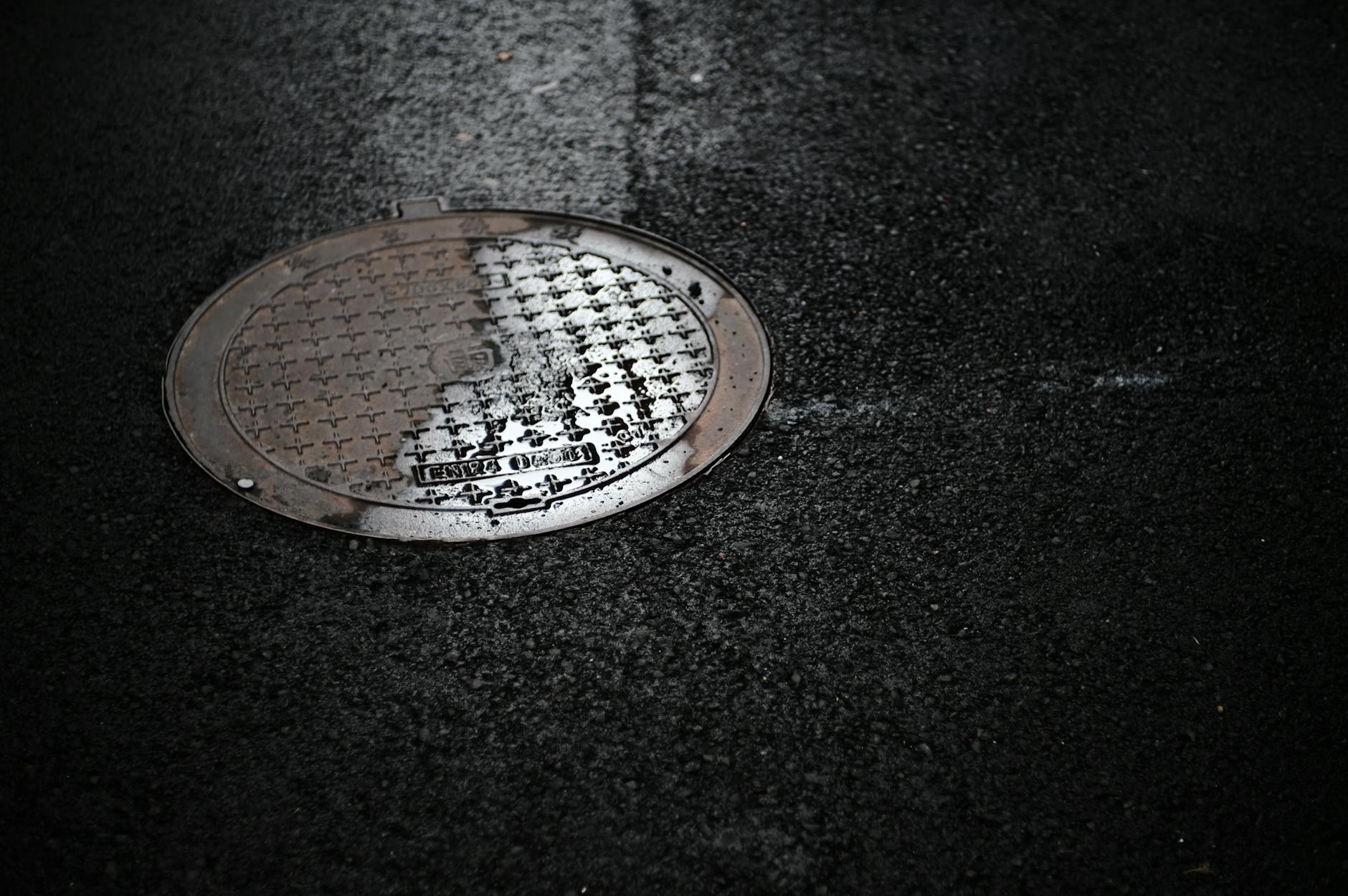 A wet manhole cover on a dark asphalt surface, reflecting light.