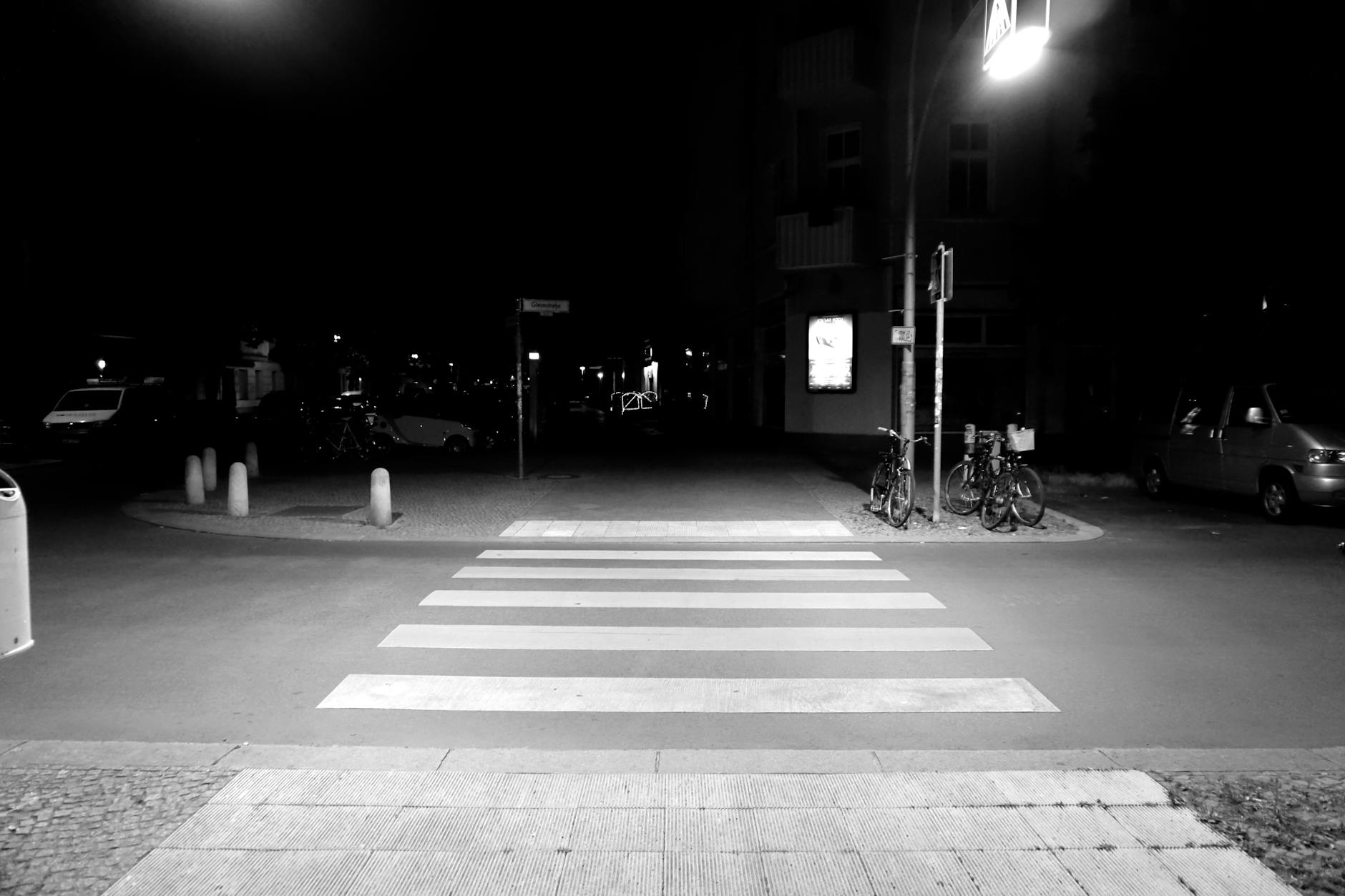 A nighttime street scene featuring a crosswalk with white stripes, surrounded by parked bicycles and vehicles. The lighting is dim, highlighting the crosswalk and nearby street signs.